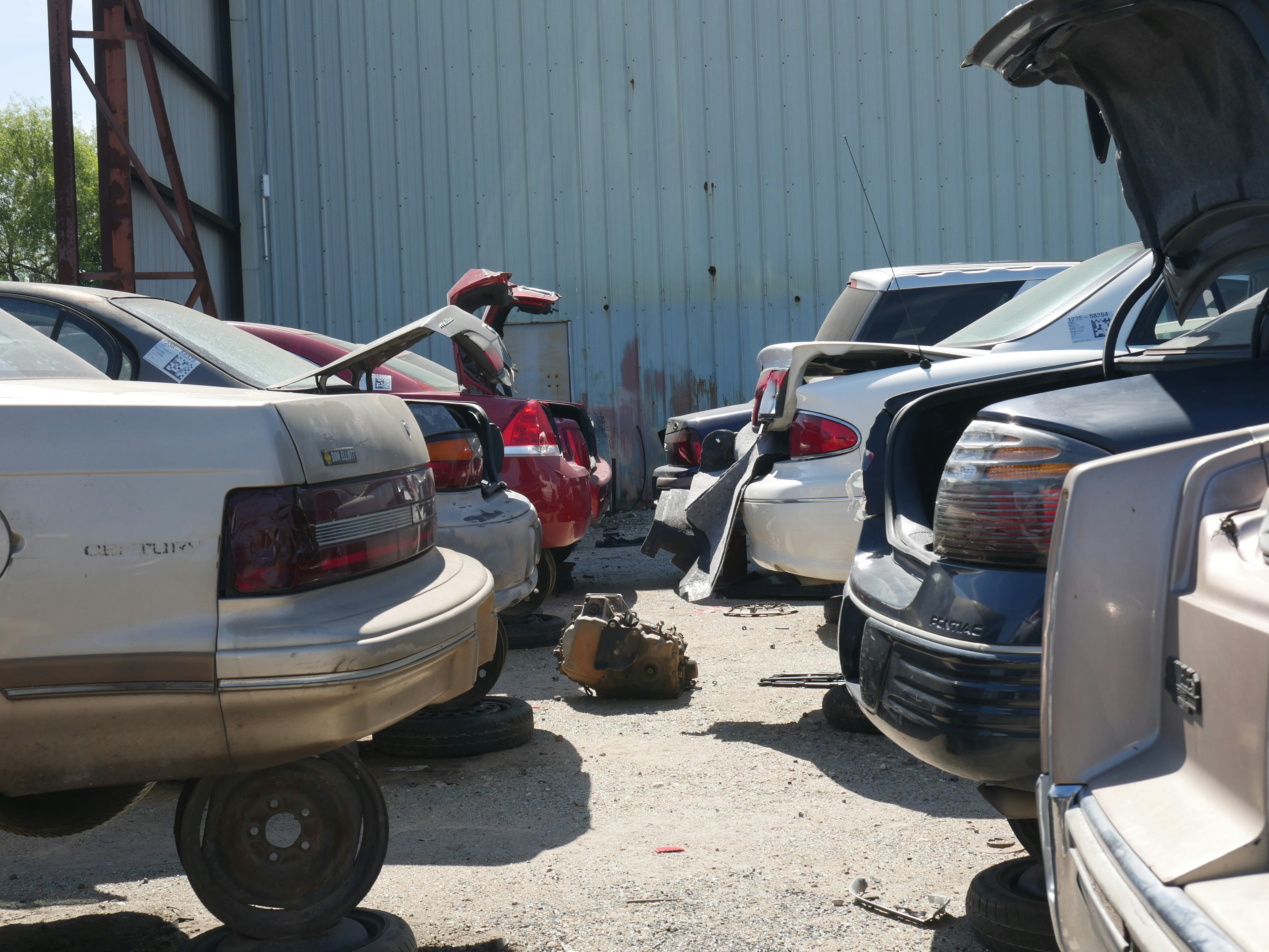 A row of cars is parked closely together in a lot, with some appearing to be missing tires, indicating a scrapyard or salvage yard setting. The cars are positioned in front of a large, corrugated metal building. Some trunks are open, and a transmission or automotive part lies on the ground.