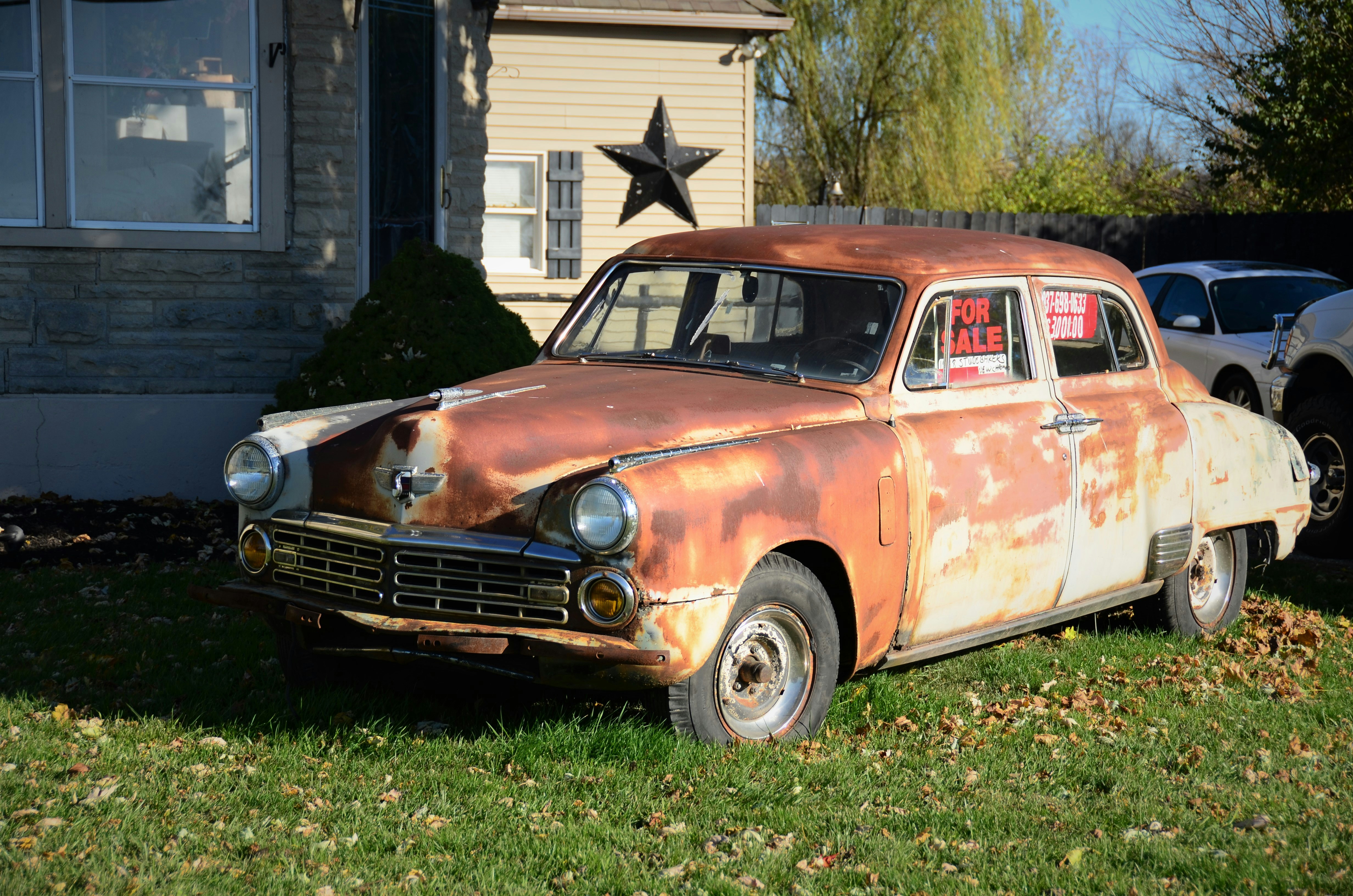 A vintage, rusty car is parked on a lawn in front of a house. The car, showing significant wear and rust, is displayed with a 'For Sale' sign in its window. The house in the background is made of stone and wood, with a large decorative star mounted on the wall. The scene is set in a daytime environment with shadows indicating bright sunlight.